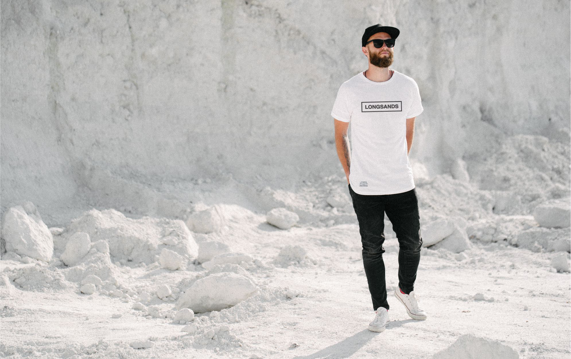 A man models the Longsands SS/24 line in a white quarry. He wears a black cap, sunglasses, the Longsands Box Tee, black jeans, and white sneakers